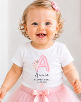 A smiling baby girl wearing a white t-shirt with a pink letter "A" and a pink tutu skirt, with a pink bow in her hair.