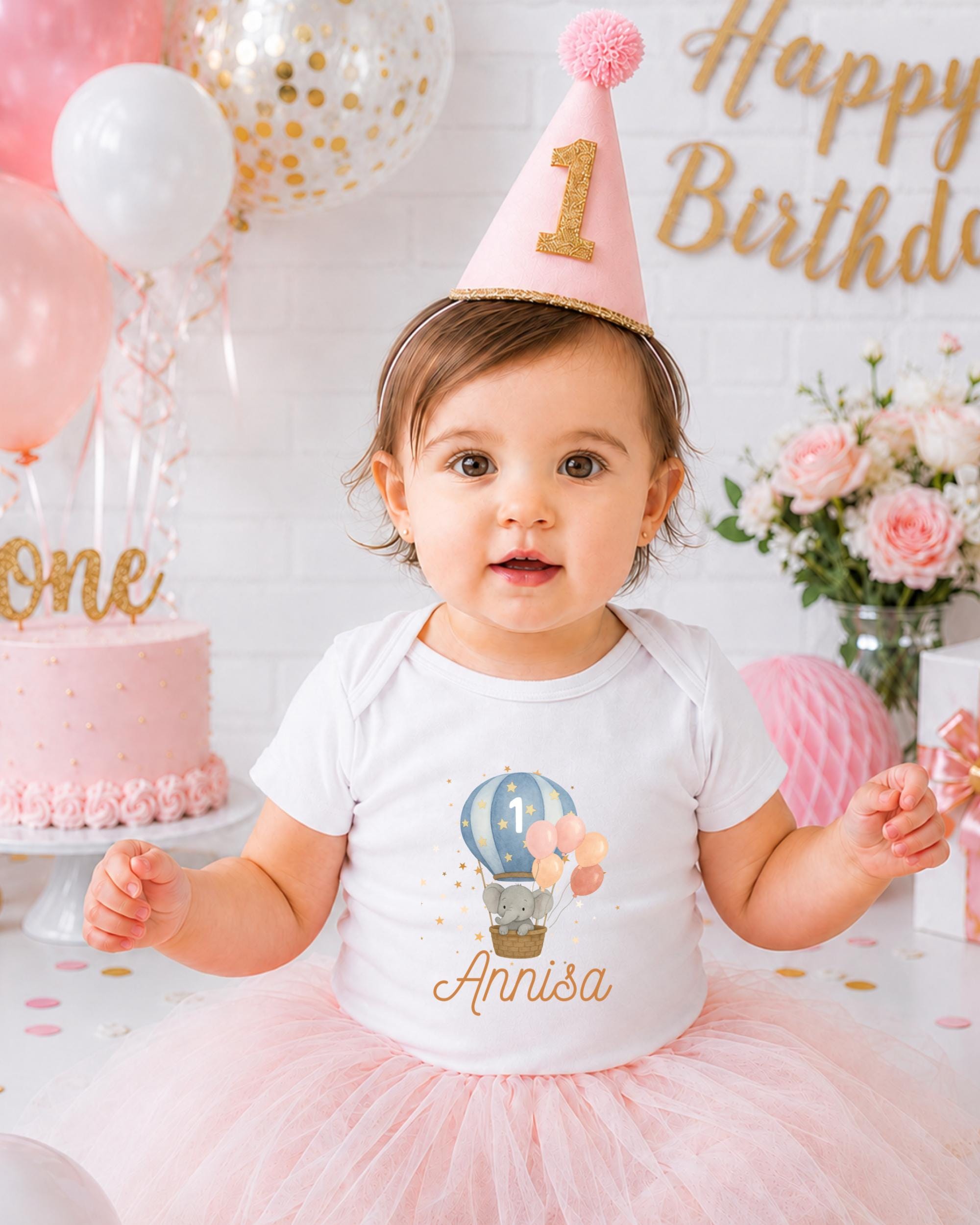 A young girl wearing a birthday hat and a white t-shirt with a balloon design is posing for a photo at a birthday party.