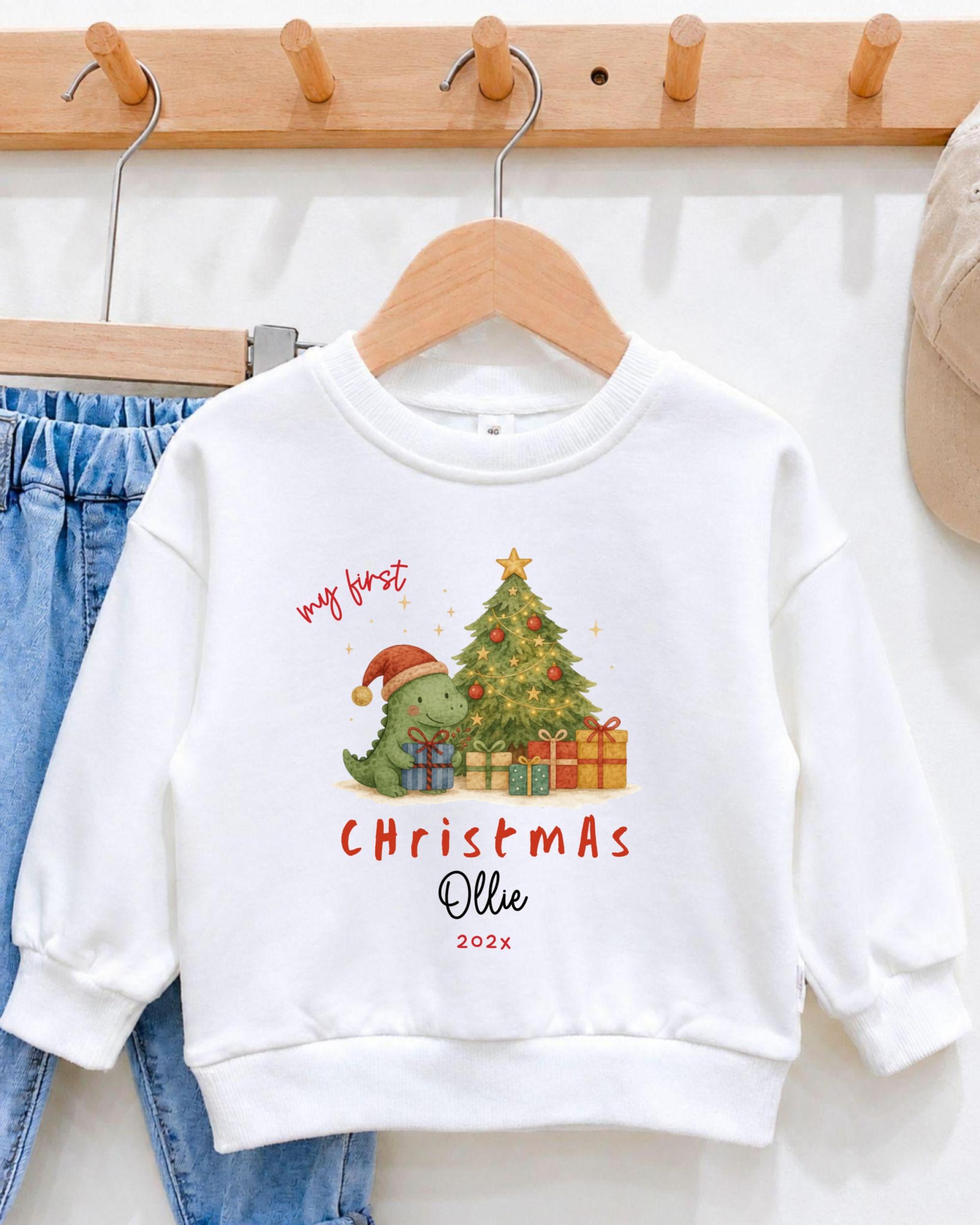A white sweatshirt with a Christmas tree and presents, along with a dinosaur wearing a Santa hat, is hanging on a wooden hanger.