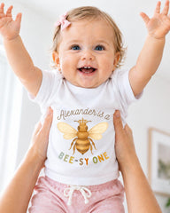 A smiling baby girl wearing a white t-shirt with a bee graphic and the text "Alexander is a BEE-SY ONE" is being held up by someone's arms.