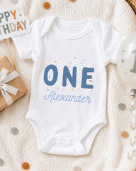 A white onesie with the text "ONE Alexander" printed on it, surrounded by confetti and a birthday sign.
