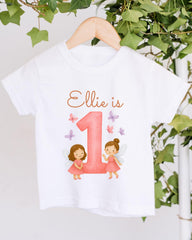 A white t-shirt with a pink number one and two-legged fairy characters printed on it, along with the text "Ellie is 1".