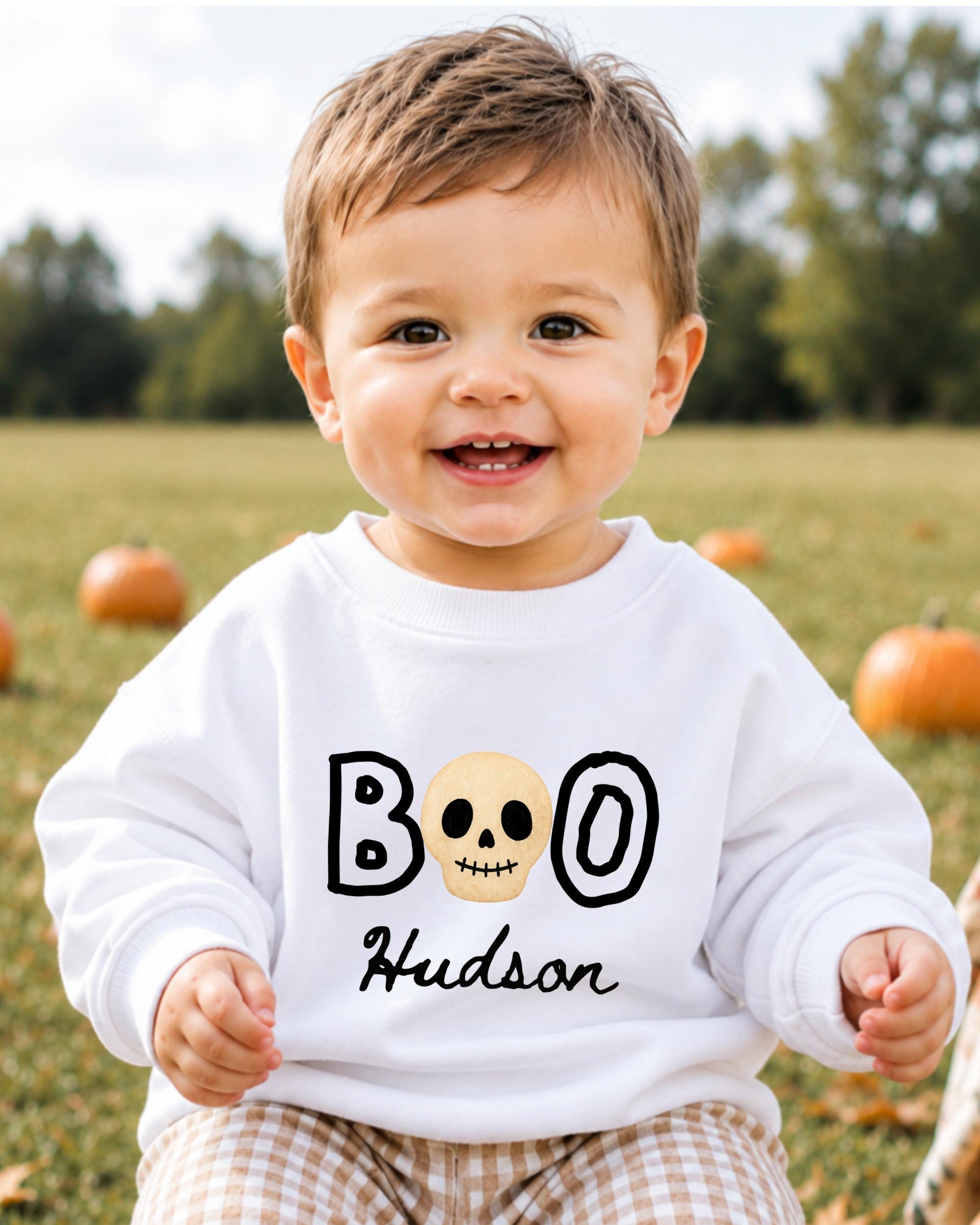 A smiling baby boy wearing a white sweater with the text "BOO Hudson" printed on it, sitting in a field with pumpkins in the background.