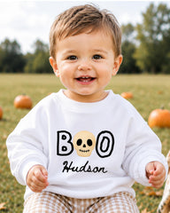 A smiling baby boy wearing a white sweater with the text "BOO Hudson" printed on it, sitting in a field with pumpkins in the background.