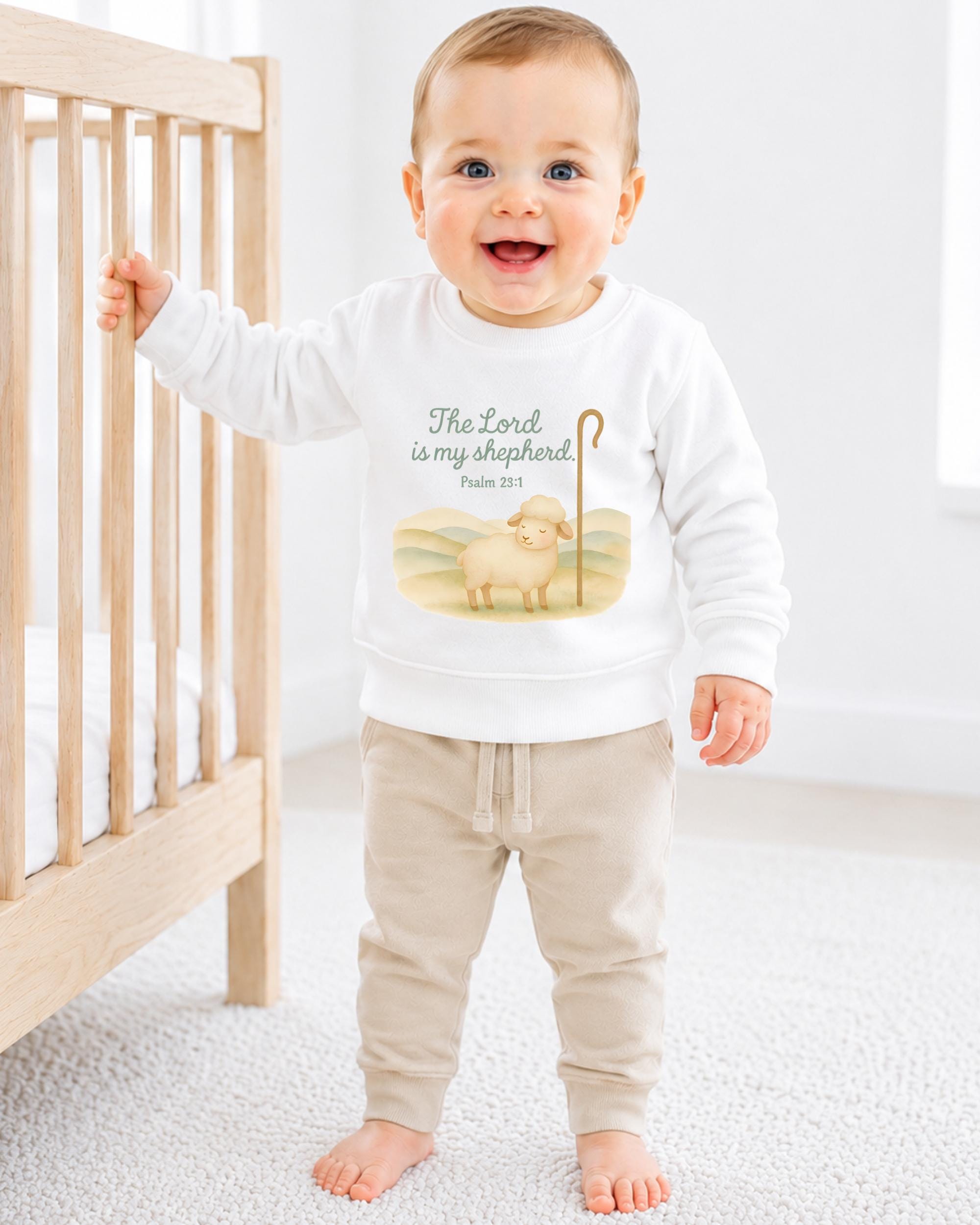 A smiling baby boy wearing a white sweatshirt with a Bible verse and a sheep graphic, standing in front of a wooden crib.