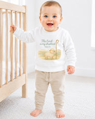 A smiling baby boy wearing a white sweatshirt with a Bible verse and a sheep graphic, standing in front of a wooden crib.