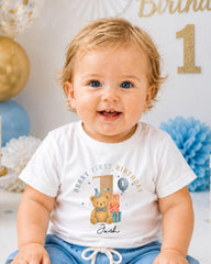 A smiling baby boy wearing a white t-shirt with a teddy bear and the text "Beany first birthday Jack" printed on it.
