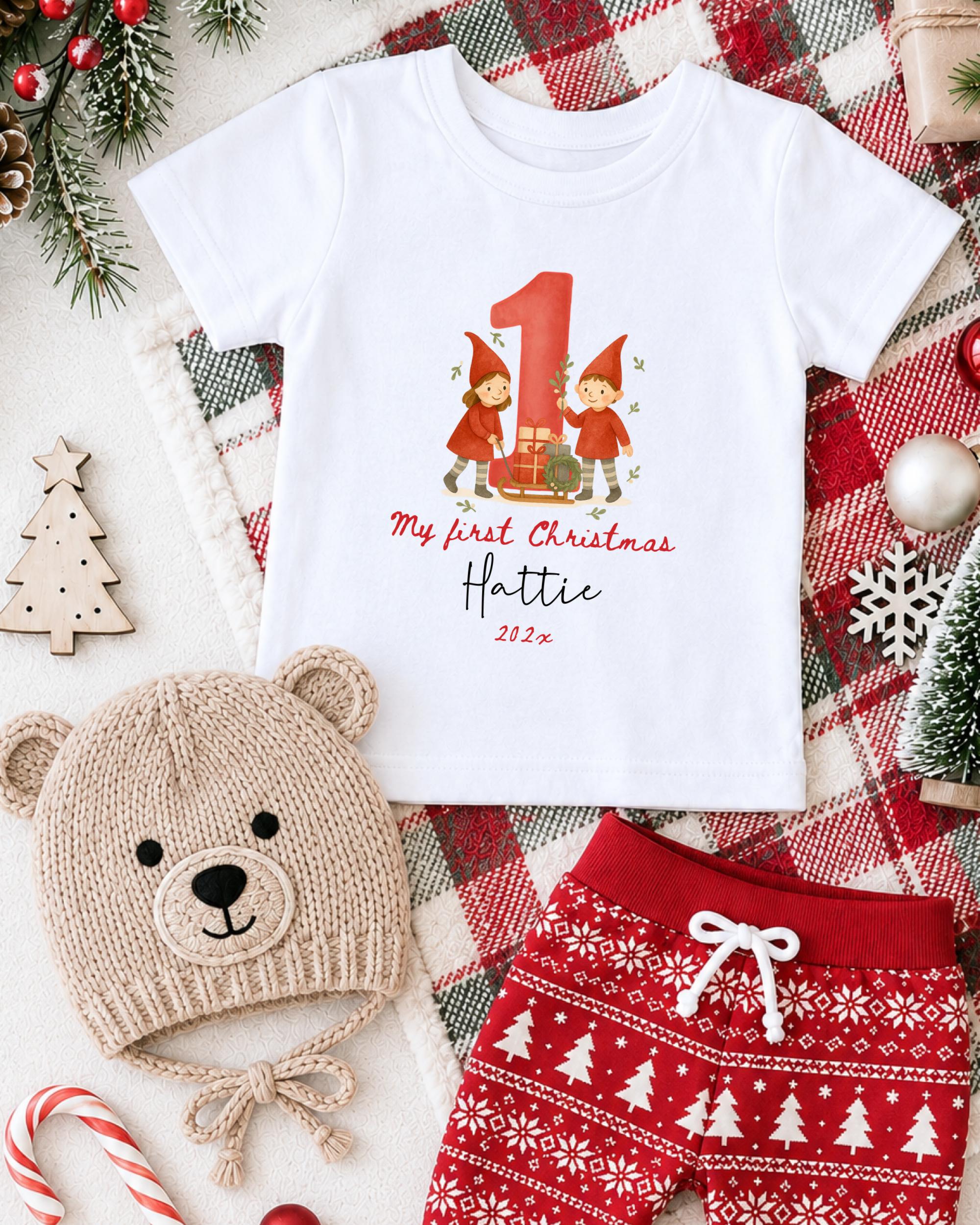 A white t-shirt with a graphic of two gnomes and the text "My first Christmas" printed on it, along with a teddy bear hat and red shorts, all set against a Christmas-themed background.