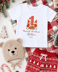 A white t-shirt with a graphic of two gnomes and the text "My first Christmas" printed on it, along with a teddy bear hat and red shorts, all set against a Christmas-themed background.