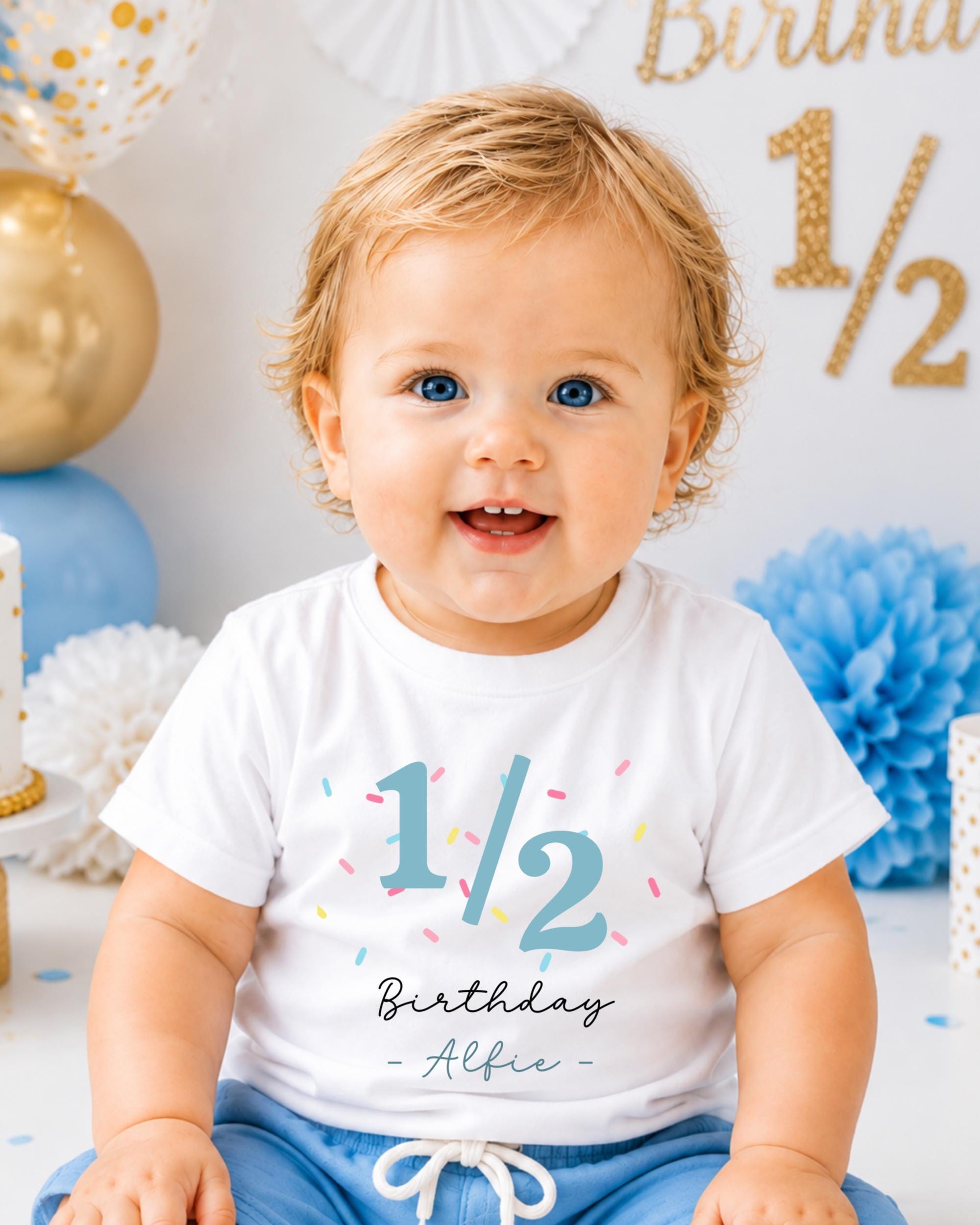 A smiling baby boy wearing a white t-shirt with the number 1/2 and the text "Birthday - Albie" printed on it, sitting in front of a birthday party backdrop with balloons and confetti.
