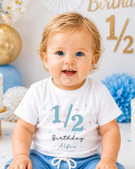 A smiling baby boy wearing a white t-shirt with the number 1/2 and the text "Birthday - Albie" printed on it, sitting in front of a birthday party backdrop with balloons and confetti.