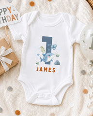 A white onesie with a shark design and the name "James" printed on it, along with a "Happy Birthday" sign and a balloon, all placed on a white blanket with confetti.