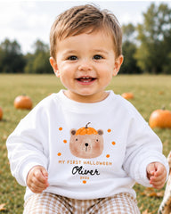 A smiling baby boy wearing a white sweatshirt with a pumpkin and bear design, sitting in a field with pumpkins in the background.