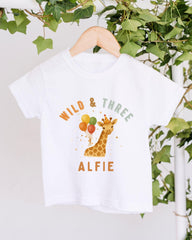 A white t-shirt with a giraffe and balloons printed on it, hanging on a wooden hanger.