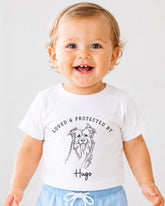 A smiling baby wearing a white t-shirt with a graphic of a dog and the text "LOVED & PROTECTED BY Hugo" printed on it.