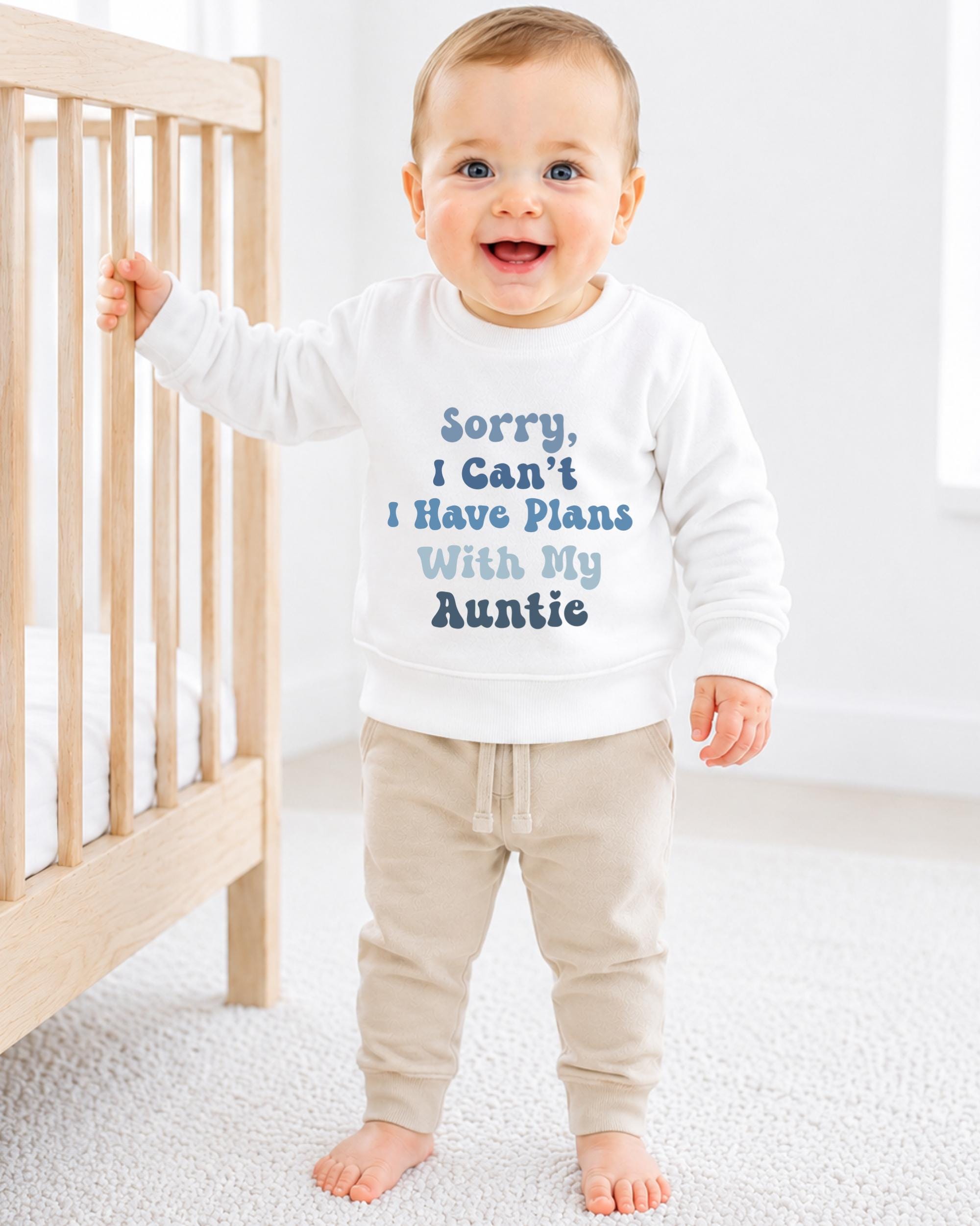 A smiling baby boy wearing a white sweatshirt with text on it, standing next to a wooden crib.
