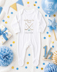 A white onesie with the text "Halfway to one Oliver" printed on it, surrounded by various baby-related items such as a blue ribbon, a star, confetti, and a balloon.