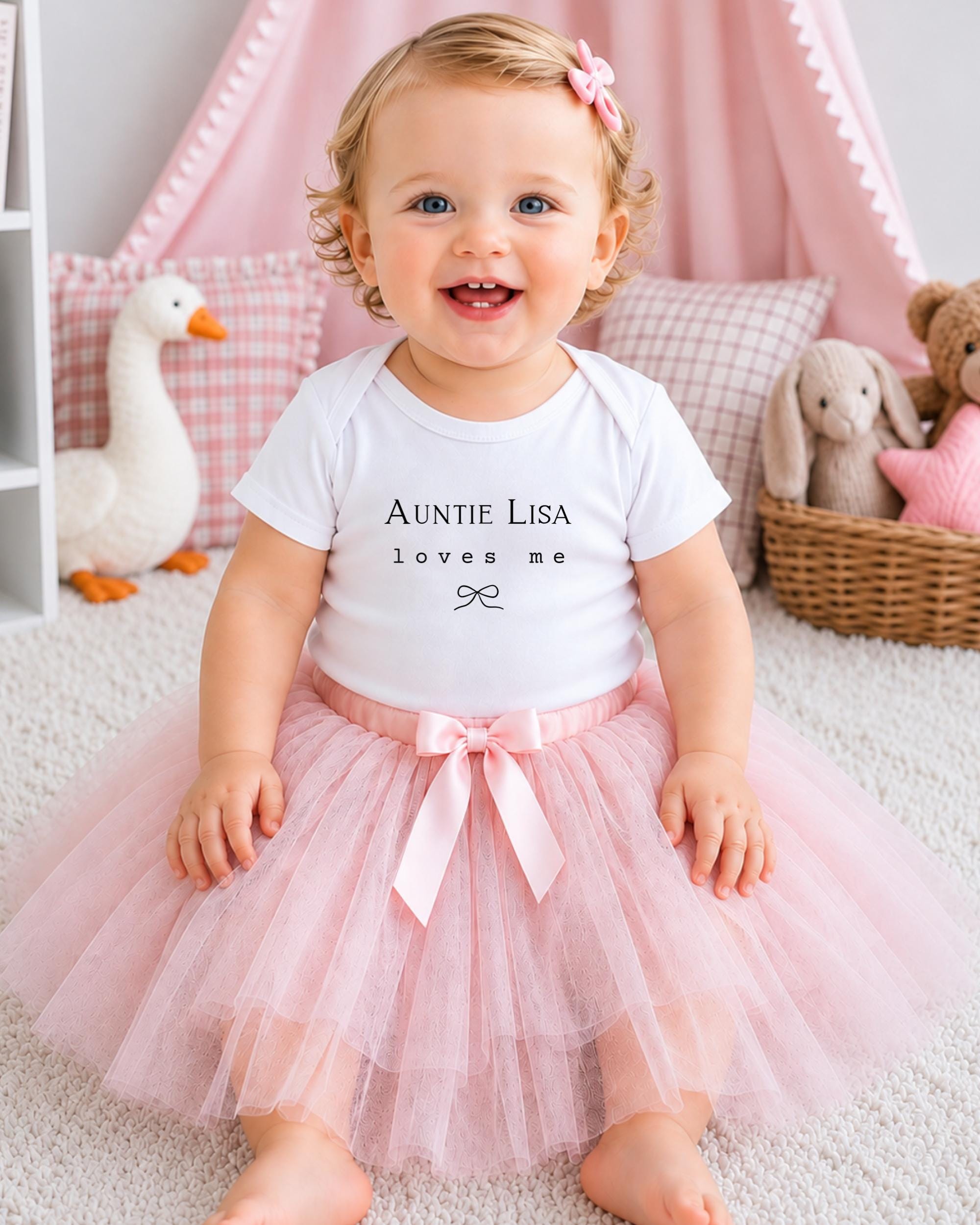 A smiling baby girl wearing a white t-shirt with the text "Auntie Lisa loves me" and a pink tutu skirt, sitting on a white carpet surrounded by stuffed animals.