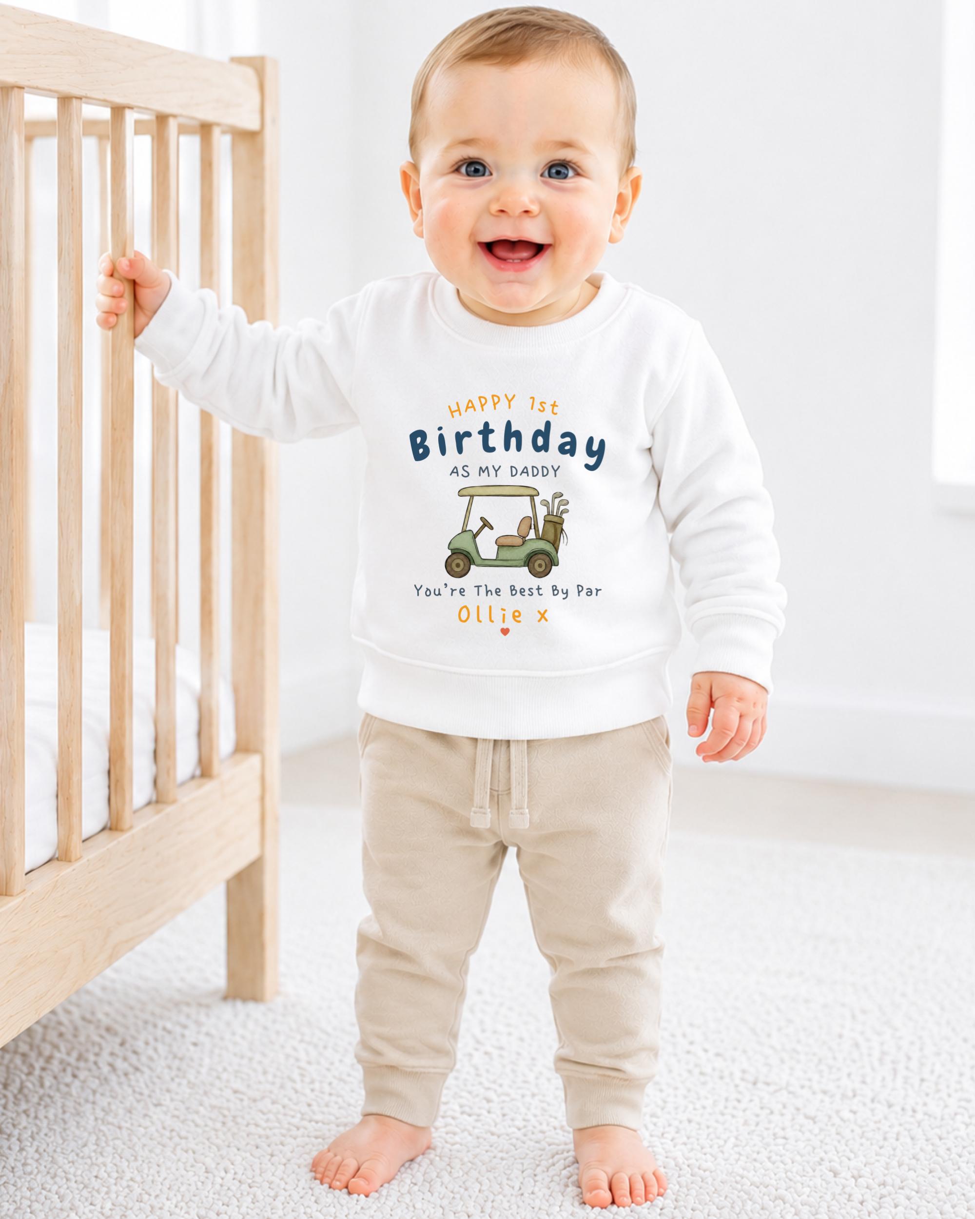 A smiling baby boy wearing a white sweatshirt with a graphic design and text on it, standing next to a wooden crib.