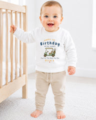 A smiling baby boy wearing a white sweatshirt with a graphic design and text on it, standing next to a wooden crib.