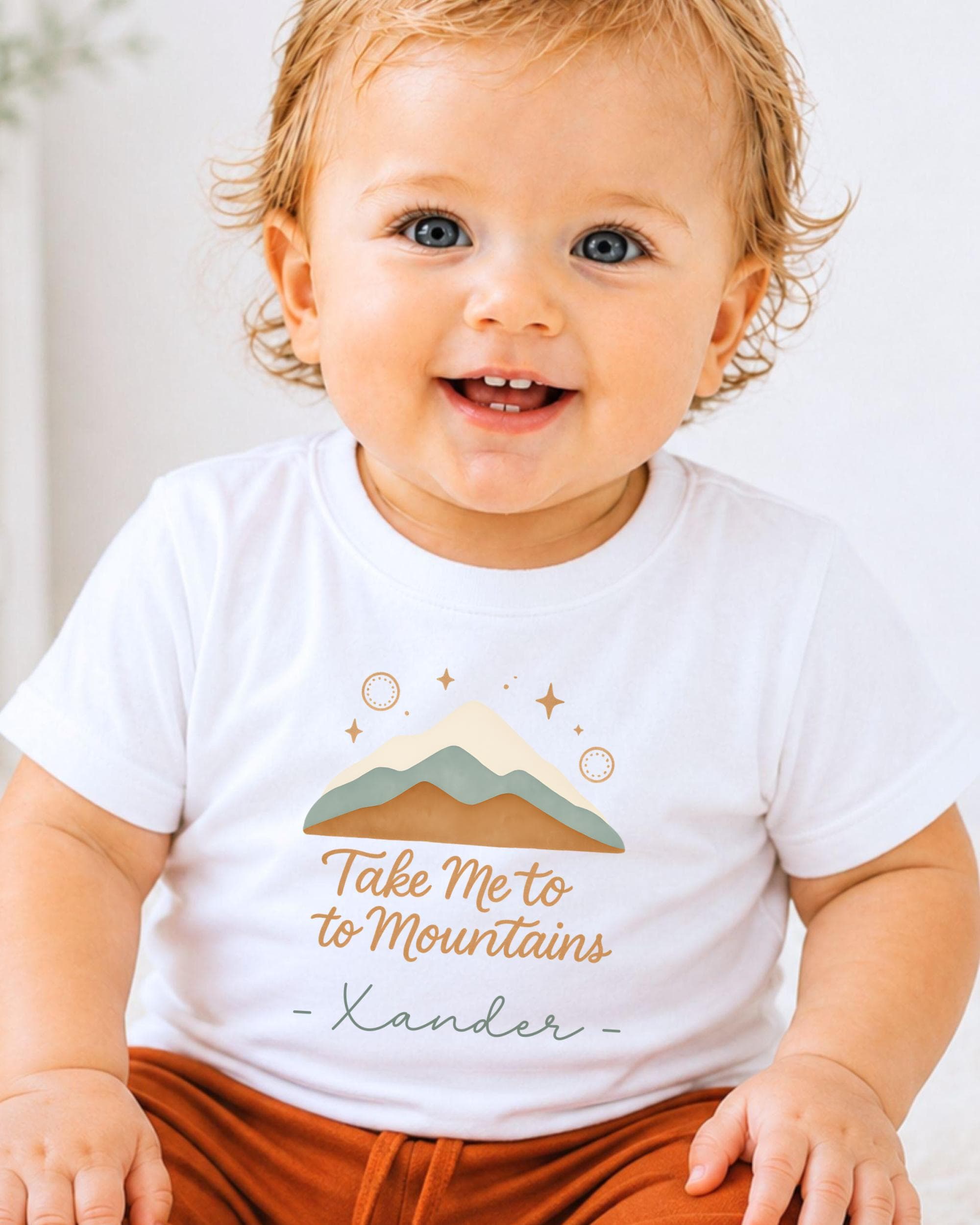 A smiling baby wearing a white t-shirt with a mountain graphic and the text "Take Me to Mountains" printed on it.