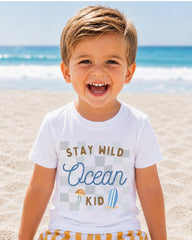 A young boy with a big smile on his face, wearing a white t-shirt with the text "STAY WILD OCEAN KID" printed on it, is standing on a sandy beach with the ocean in the background.
