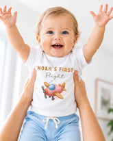 A smiling baby wearing a white t-shirt with a pink airplane graphic and the text "Noah's first flight" is being held up by two people.