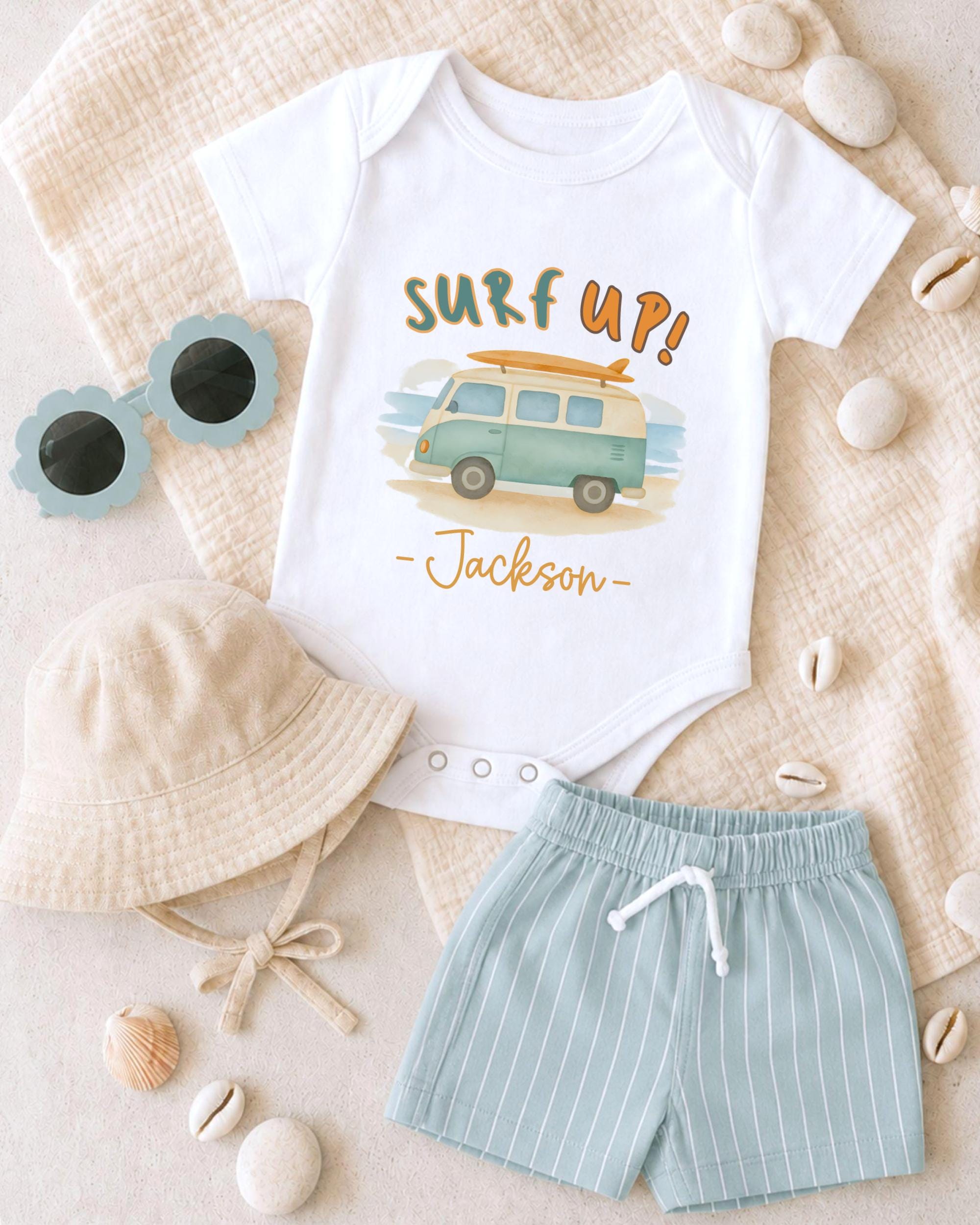 A baby's outfit, including a white onesie with a surfboard graphic, a beige bucket hat, and light blue shorts, along with sunglasses and seashells, are arranged on a beige surface.