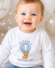 A smiling baby boy wearing a white shirt with a teddy bear and a balloon on it, celebrating his first birthday.
