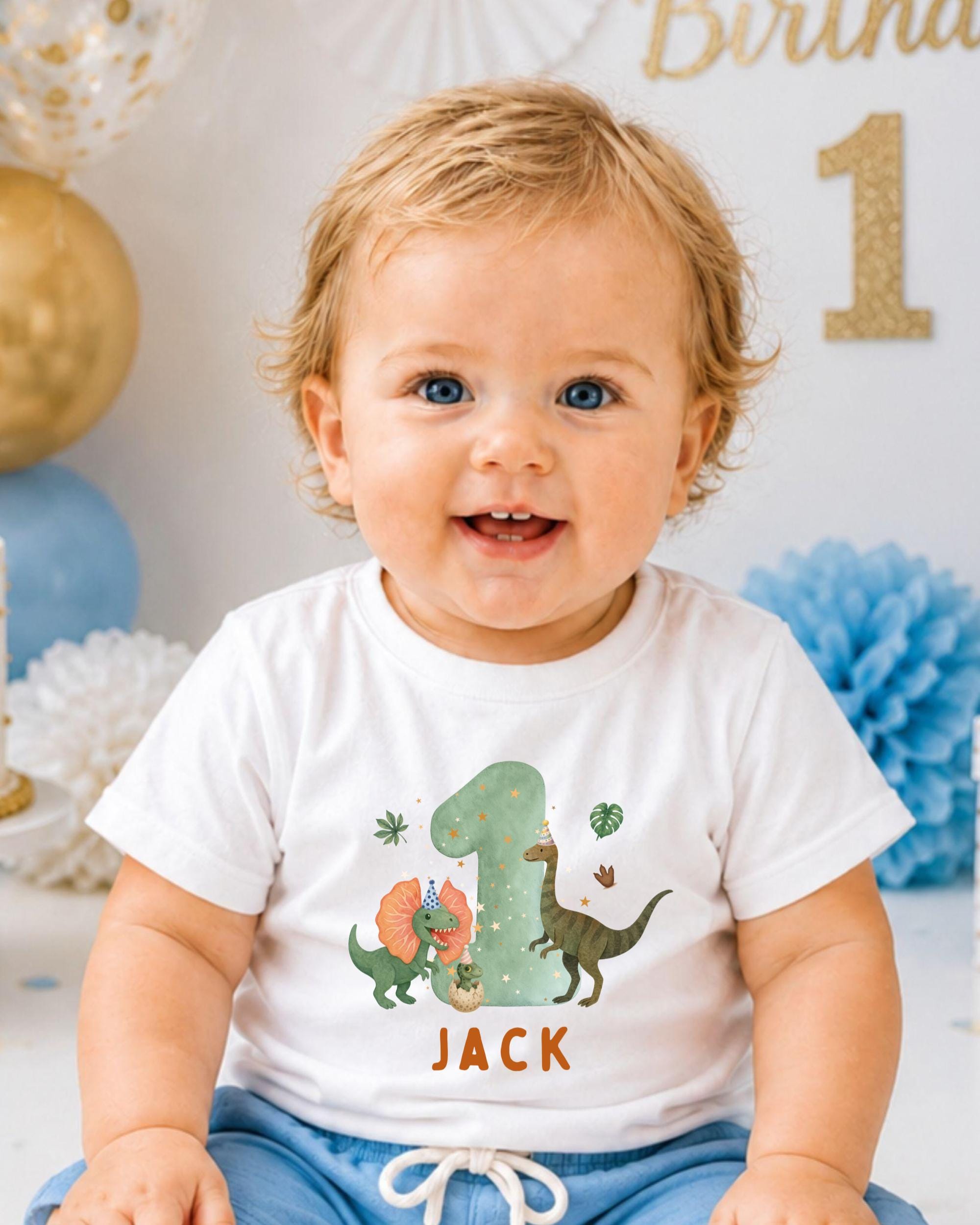 A smiling baby boy wearing a white t-shirt with a dinosaur graphic and the name 'Jack' printed on it, sitting in front of a birthday party backdrop.