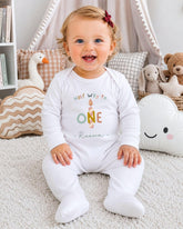 A smiling baby girl wearing a white onesie with the text "Half Way to One" sits on a white carpet surrounded by stuffed animals and pillows.