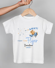 A white t-shirt with a graphic design featuring a teddy bear flying in a plane and the text "UP UP AWAY! FIRST TIME FLYER Barcelona 20XX".