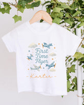 A white t-shirt with a graphic design featuring airplanes and clouds, along with the text "First Time Flyer" and the name "Kater" printed on it.