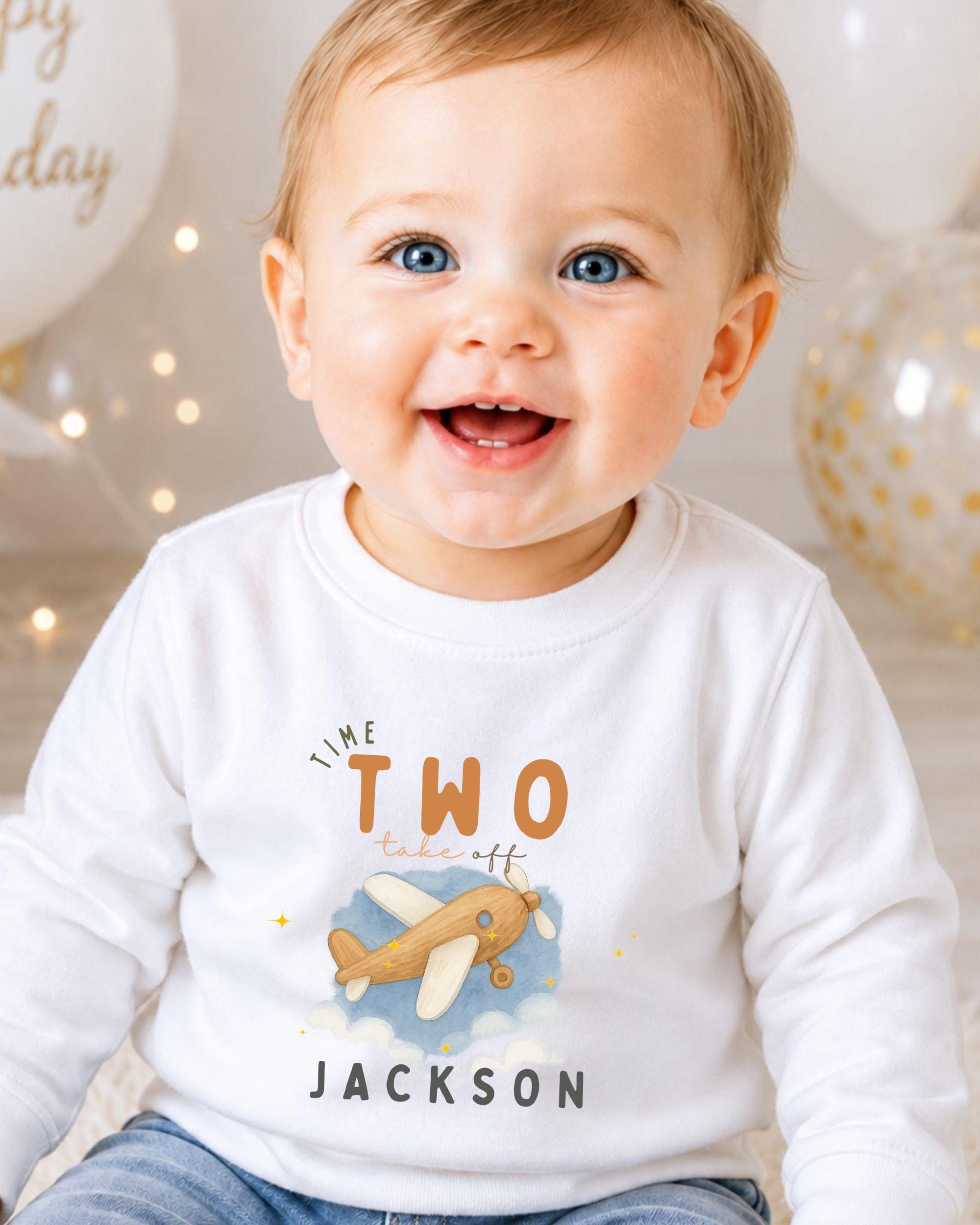 A smiling baby boy wearing a white shirt with a cartoon airplane and the text "Time Two Take Off Jackson" printed on it.