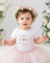 A young girl wearing a white t-shirt with a floral design and a pink tutu, posing in front of a white brick wall with flowers.