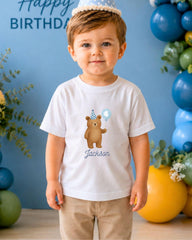 A young boy wearing a white t-shirt with a cartoon bear and the name "Jackson" printed on it, standing in front of a birthday backdrop with balloons.