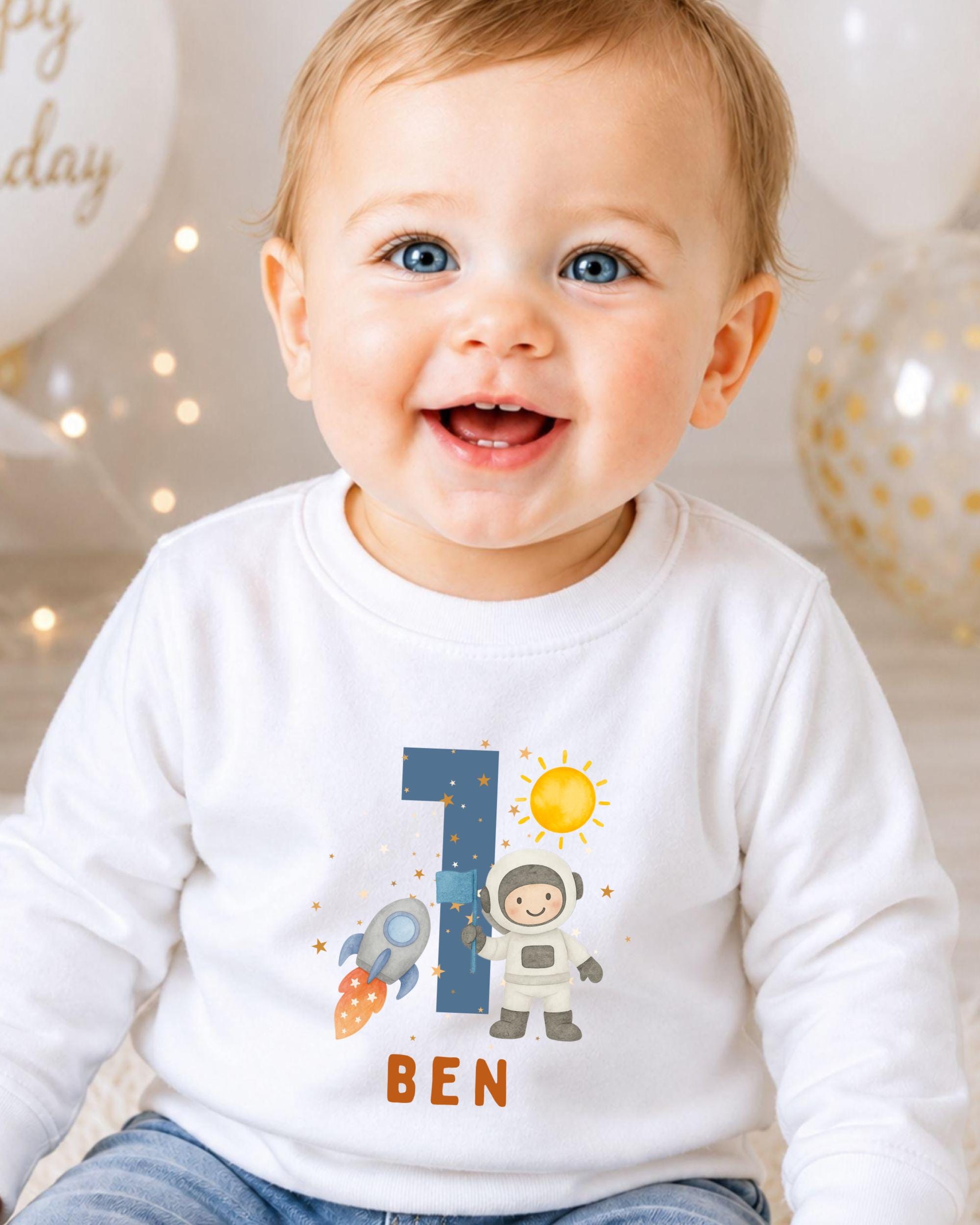 A smiling baby boy wearing a white shirt with a space-themed design and the name 'Ben' printed on it.