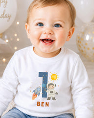 A smiling baby boy wearing a white shirt with a space-themed design and the name 'Ben' printed on it.