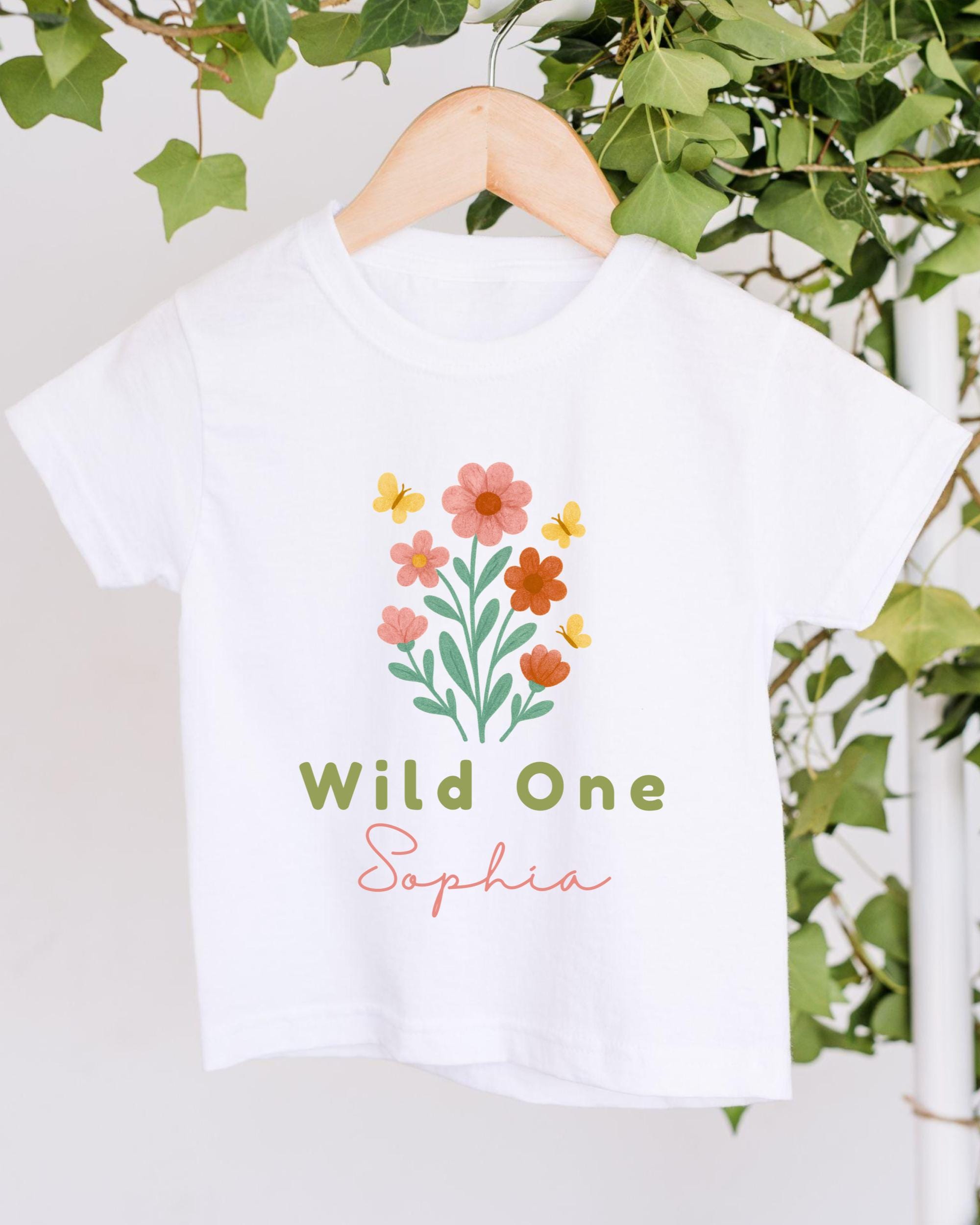 A white t-shirt with a floral design and the text "Wild One Sophia" printed on it, hanging on a wooden hanger.
