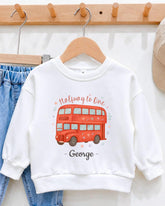 A white sweatshirt with a red double-decker bus design and the text "Halfway to One George" printed on it, hanging on a wooden hanger.
