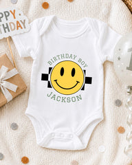 A white onesie with a yellow smiley face and the text "Birthday Boy Jackson" printed on it, along with a "Happy Birthday" sign and confetti.