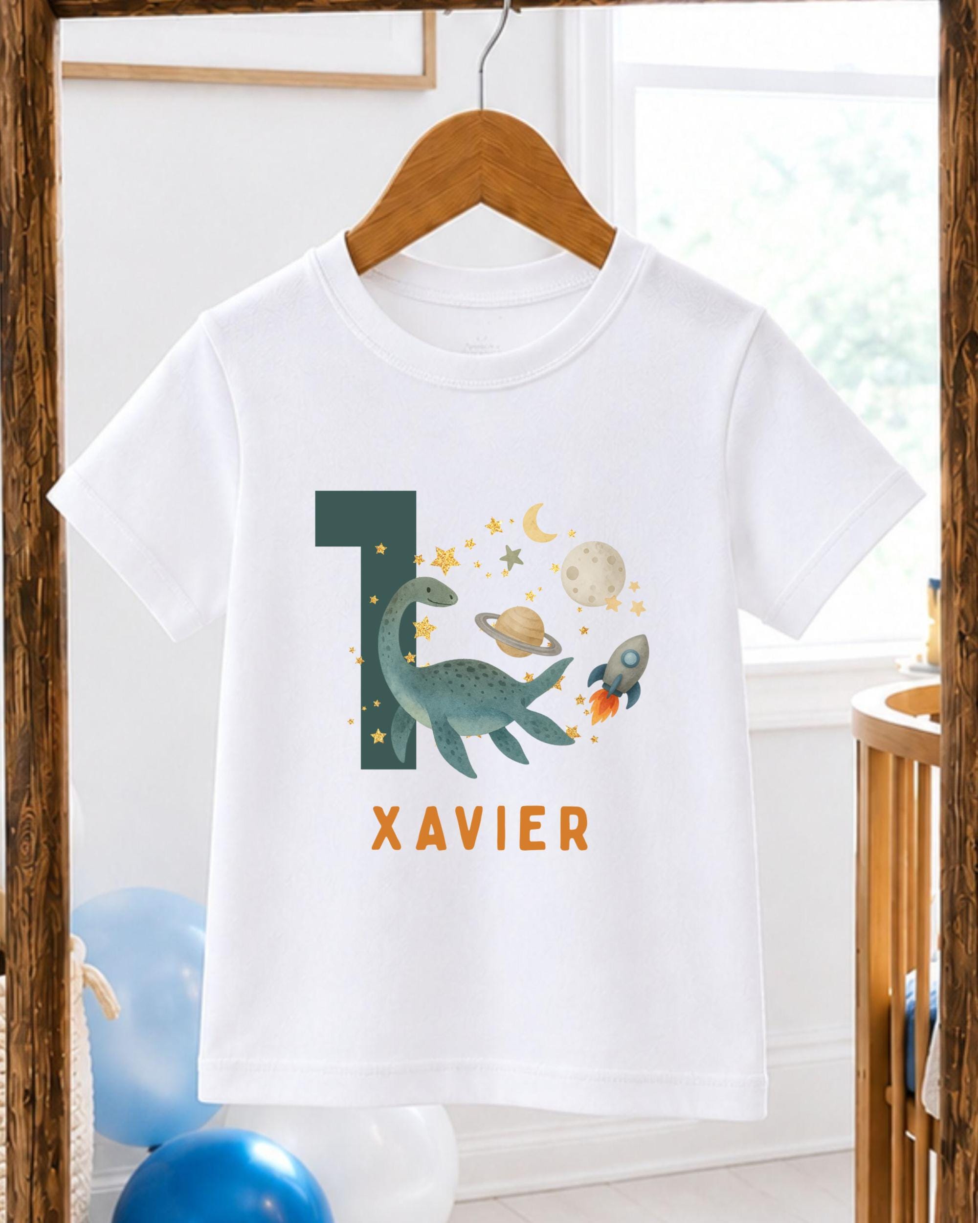 A white t-shirt with a graphic design featuring a dinosaur, a rocket ship, and a planet, along with the text "XAVIER" printed on it.