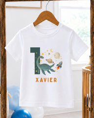 A white t-shirt with a graphic design featuring a dinosaur, a rocket ship, and a planet, along with the text "XAVIER" printed on it.
