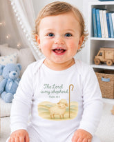 A smiling baby wearing a white onesie with a Bible verse printed on it, sitting in a room with a teddy bear and bookshelf in the background.