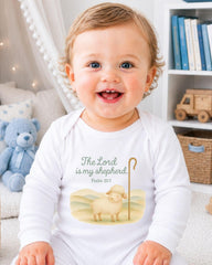 A smiling baby wearing a white onesie with a Bible verse printed on it, sitting in a room with a teddy bear and bookshelf in the background.