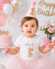 A young girl wearing a birthday hat and a white t-shirt with a giraffe design is posing in front of a birthday cake and balloons.