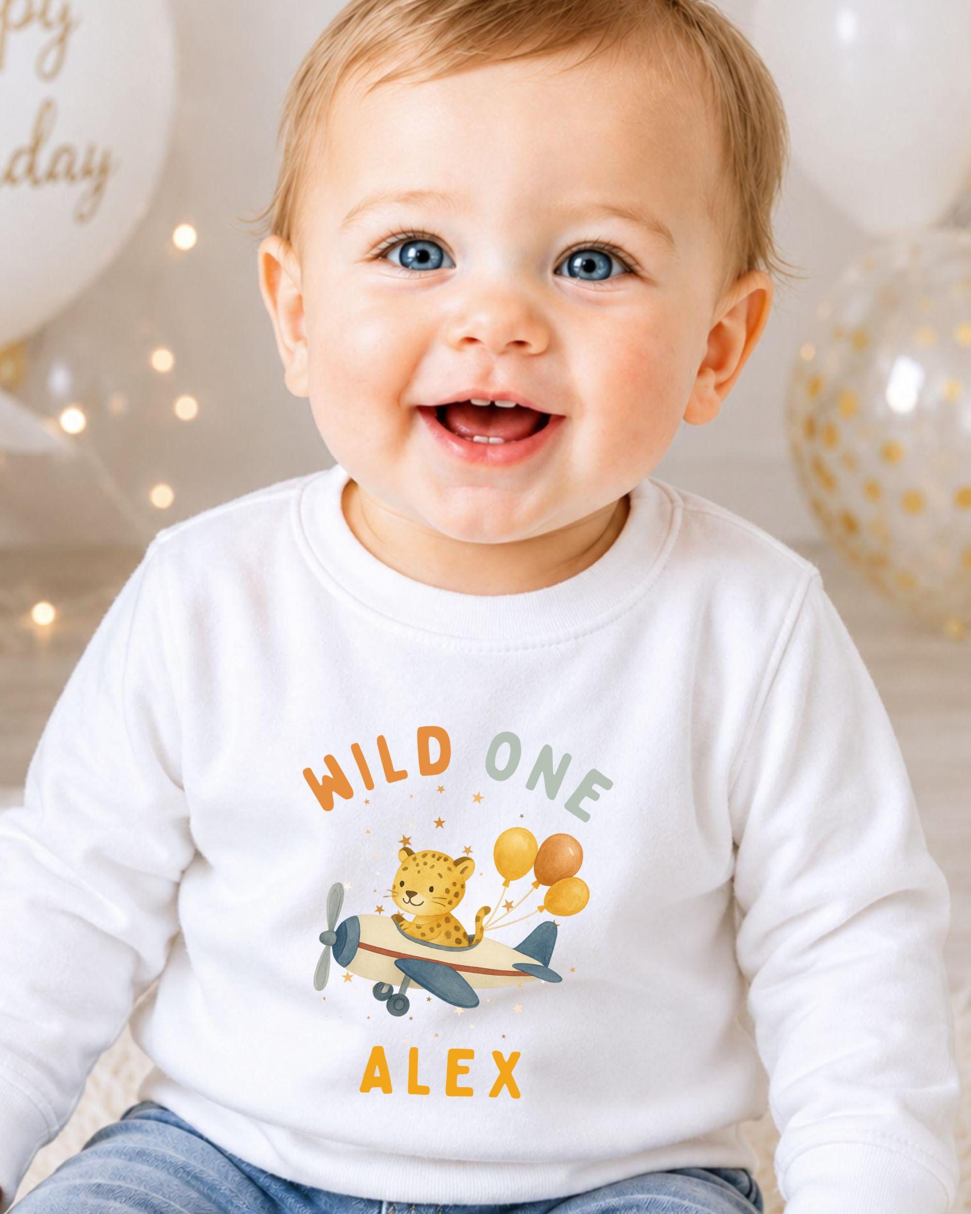 A smiling baby boy wearing a white shirt with a cartoon airplane and the text "WILD ONE ALEX" printed on it.