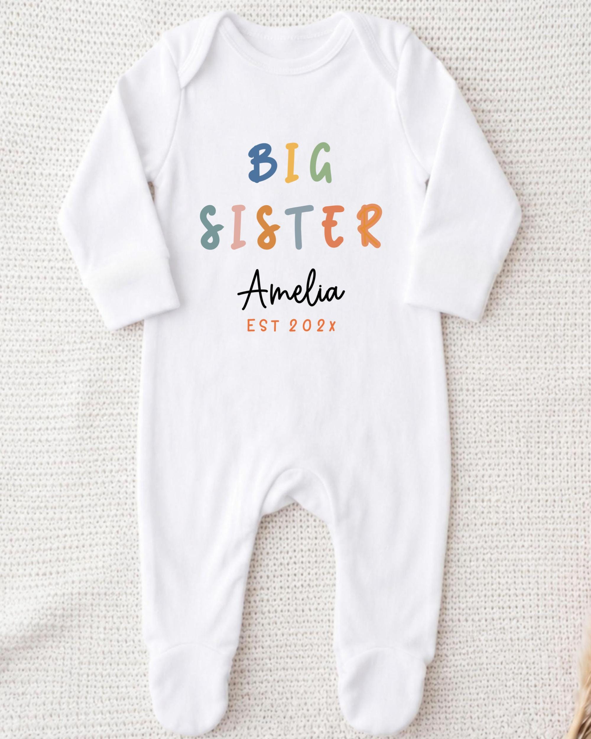 A white onesie with the text "BIG SISTER Amelia EST 202x" printed on it.
