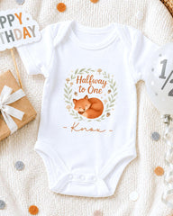 A white onesie with a cartoon image of a fox and the text "Halfway to One" printed on it, along with the name "Knox" below the image. The onesie is placed on a white blanket, surrounded by various party decorations such as a birthday sign, a balloon, and confetti.