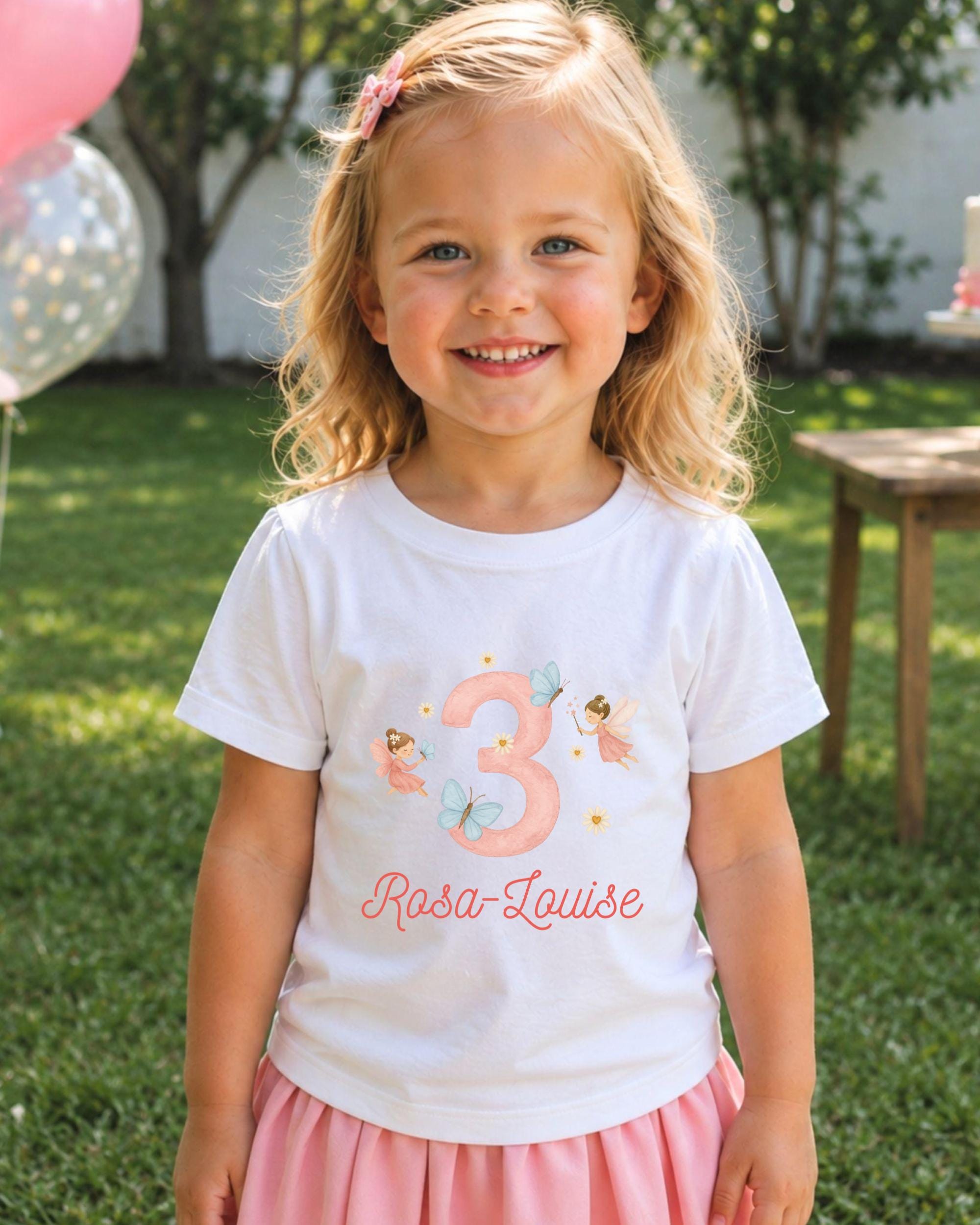 A young girl with blonde hair is smiling and wearing a white t-shirt with a pink number 3 and a fairy design, standing in a grassy area with balloons in the background.
